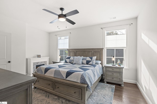 bedroom with a tiled fireplace, dark wood-type flooring, multiple windows, and ceiling fan