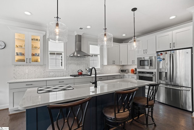 kitchen with appliances with stainless steel finishes, a kitchen island with sink, wall chimney exhaust hood, and hanging light fixtures