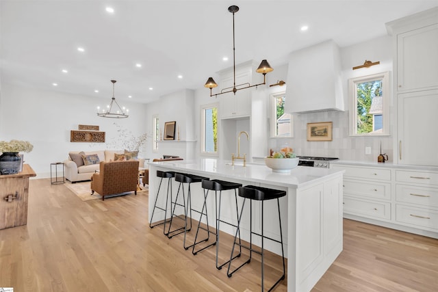 kitchen featuring a large island with sink, custom range hood, decorative light fixtures, white cabinets, and light wood-type flooring