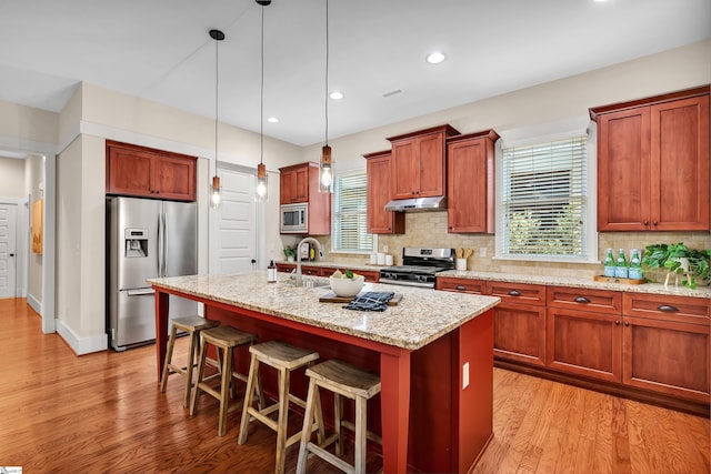 kitchen with stainless steel appliances, decorative light fixtures, light stone counters, a kitchen island with sink, and a breakfast bar