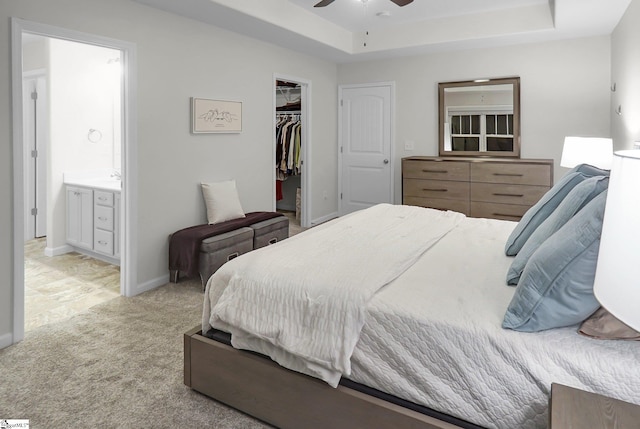 carpeted bedroom with a tray ceiling, a walk in closet, ensuite bathroom, and ceiling fan