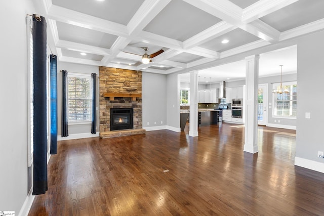 unfurnished living room with dark hardwood / wood-style flooring, ornate columns, ceiling fan with notable chandelier, beam ceiling, and a stone fireplace