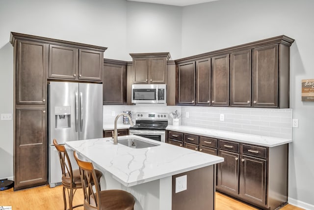 kitchen with appliances with stainless steel finishes, tasteful backsplash, light stone counters, light hardwood / wood-style flooring, and an island with sink