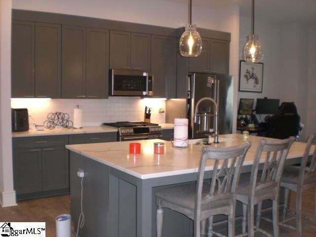 kitchen featuring hanging light fixtures, stainless steel appliances, backsplash, gray cabinets, and a kitchen island with sink