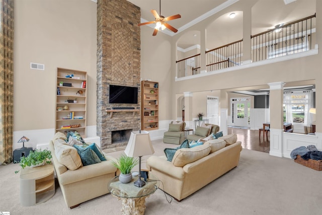 carpeted living room featuring a stone fireplace, built in shelves, ceiling fan, a towering ceiling, and ornate columns