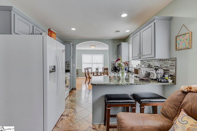 kitchen with tasteful backsplash, white refrigerator with ice dispenser, kitchen peninsula, dark stone counters, and gray cabinets