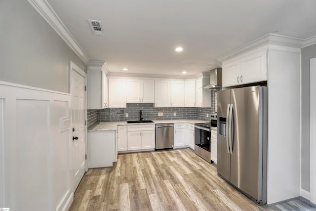 kitchen with appliances with stainless steel finishes, light wood-type flooring, ornamental molding, wall chimney range hood, and white cabinets