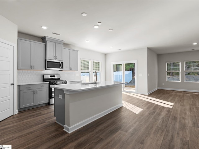 kitchen with dark wood-type flooring, stainless steel appliances, an island with sink, gray cabinets, and decorative backsplash