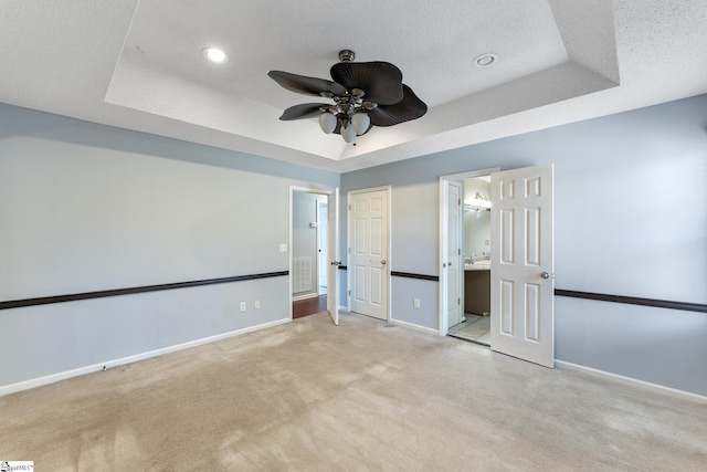 unfurnished bedroom with light carpet, ensuite bathroom, a textured ceiling, a tray ceiling, and ceiling fan