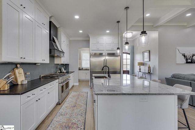 kitchen with high quality appliances, a center island with sink, decorative light fixtures, and wall chimney range hood