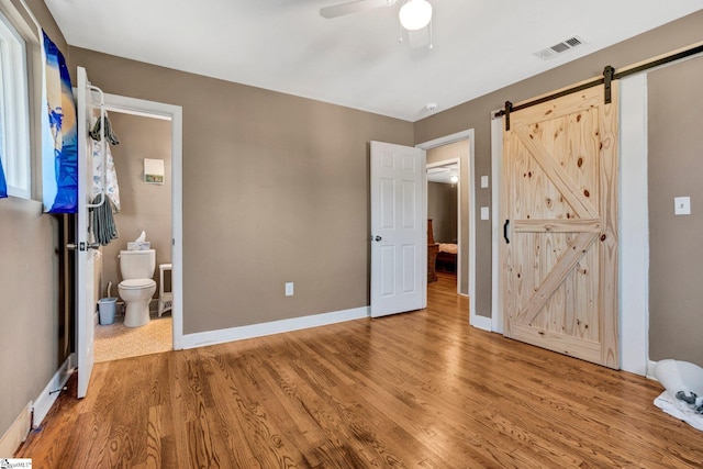 unfurnished bedroom with hardwood / wood-style floors, a barn door, ensuite bath, and ceiling fan