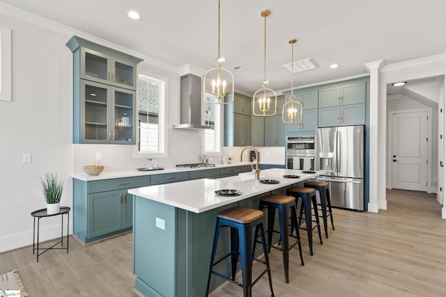 kitchen featuring a kitchen island with sink, wall chimney range hood, hanging light fixtures, light wood-type flooring, and appliances with stainless steel finishes