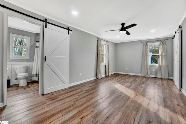 unfurnished bedroom with light wood-type flooring, ceiling fan, a barn door, multiple windows, and connected bathroom