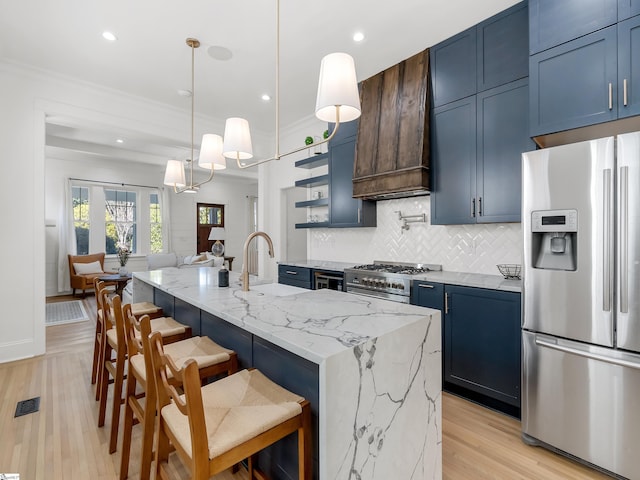 kitchen featuring stainless steel appliances, tasteful backsplash, light stone counters, a center island with sink, and light wood-type flooring