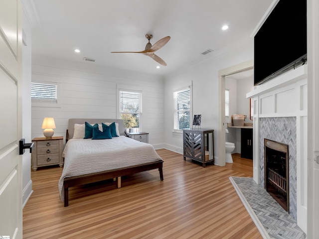 bedroom featuring connected bathroom, a tiled fireplace, ceiling fan, and light wood-type flooring