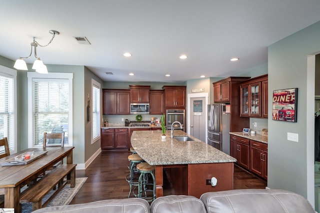 kitchen featuring dark wood-type flooring, a center island with sink, sink, appliances with stainless steel finishes, and light stone counters