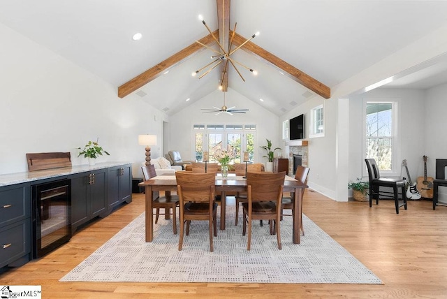 dining room featuring lofted ceiling with beams, ceiling fan with notable chandelier, a stone fireplace, light hardwood / wood-style floors, and beverage cooler