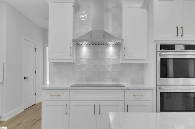 kitchen featuring black electric stovetop, wall chimney exhaust hood, stainless steel double oven, white cabinets, and light hardwood / wood-style floors