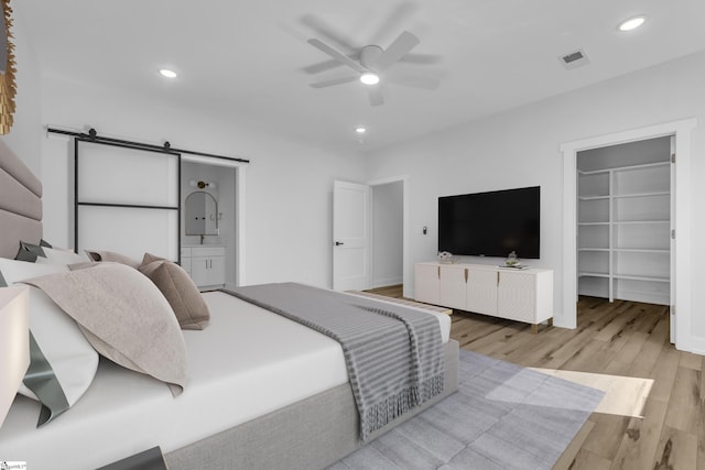 bedroom with a walk in closet, ensuite bath, light hardwood / wood-style flooring, ceiling fan, and a barn door