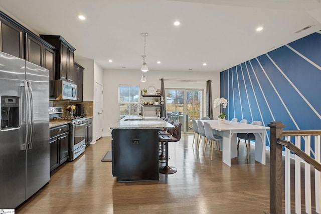 kitchen featuring stainless steel appliances, a kitchen breakfast bar, light stone counters, decorative light fixtures, and a kitchen island with sink