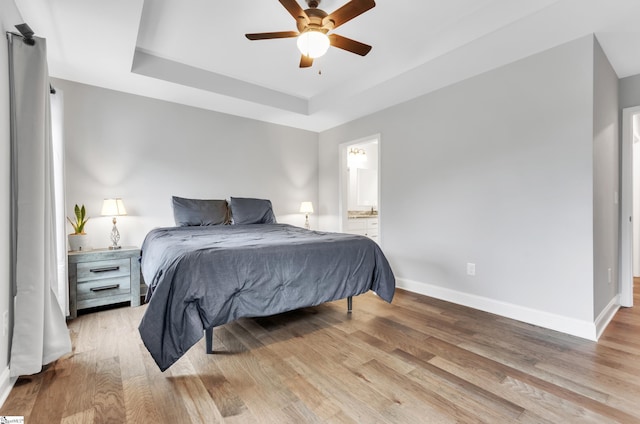 bedroom featuring a tray ceiling, ceiling fan, ensuite bathroom, and light wood-type flooring