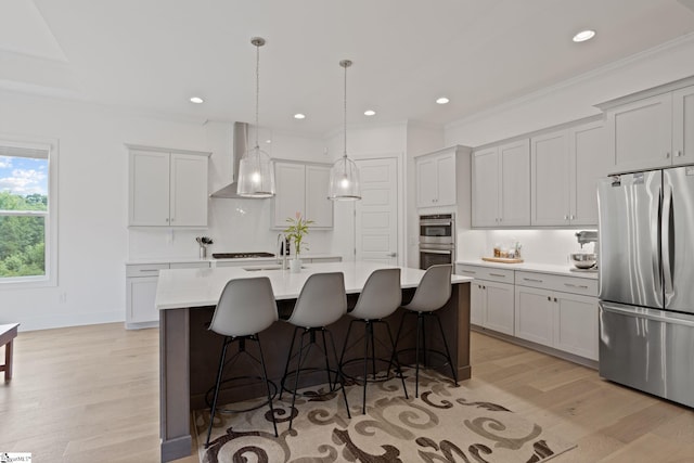 kitchen with hanging light fixtures, wall chimney range hood, light hardwood / wood-style flooring, a kitchen island with sink, and appliances with stainless steel finishes