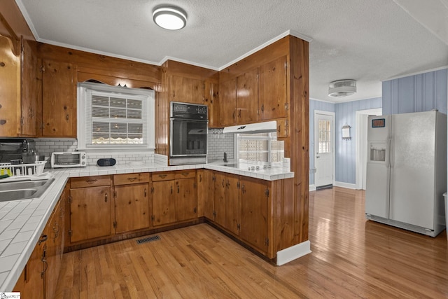 kitchen with tasteful backsplash, white fridge with ice dispenser, black oven, tile counters, and light hardwood / wood-style floors