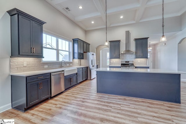 kitchen with pendant lighting, wall chimney range hood, light wood-type flooring, appliances with stainless steel finishes, and a kitchen island