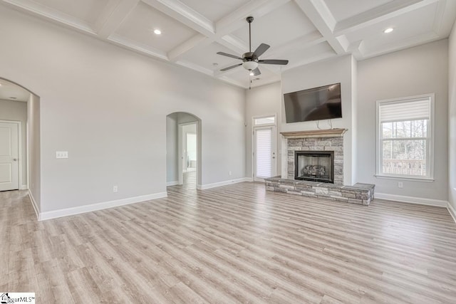 unfurnished living room featuring light wood-type flooring, coffered ceiling, ceiling fan, beam ceiling, and a stone fireplace