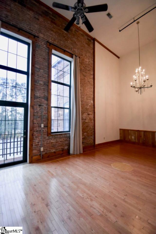 empty room featuring wood-type flooring, ceiling fan with notable chandelier, a high ceiling, and brick wall