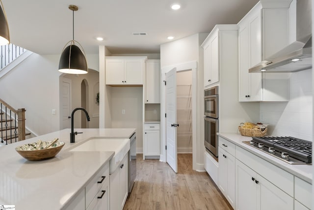 kitchen with wall chimney range hood, white cabinetry, hanging light fixtures, light wood-type flooring, and stainless steel appliances