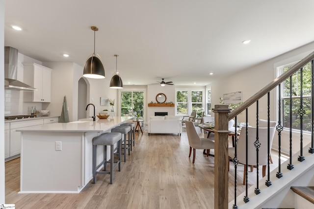 kitchen featuring decorative light fixtures, a large fireplace, an island with sink, white cabinets, and wall chimney exhaust hood