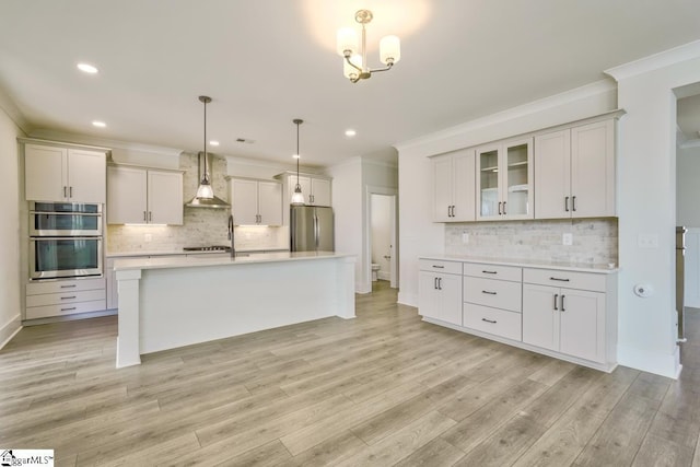 kitchen with a kitchen island with sink, white cabinets, hanging light fixtures, wall chimney exhaust hood, and appliances with stainless steel finishes