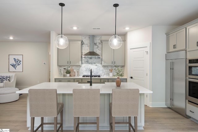 kitchen with a center island with sink, appliances with stainless steel finishes, wall chimney range hood, hanging light fixtures, and decorative backsplash