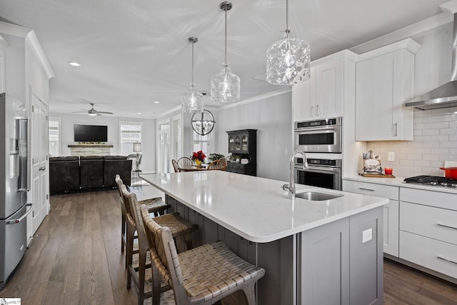 kitchen featuring pendant lighting, wall chimney range hood, decorative backsplash, an island with sink, and appliances with stainless steel finishes