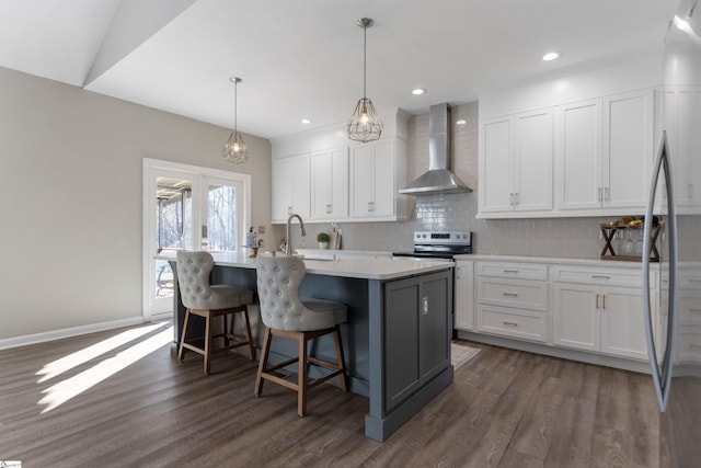 kitchen with white cabinetry, wall chimney range hood, an island with sink, and hanging light fixtures