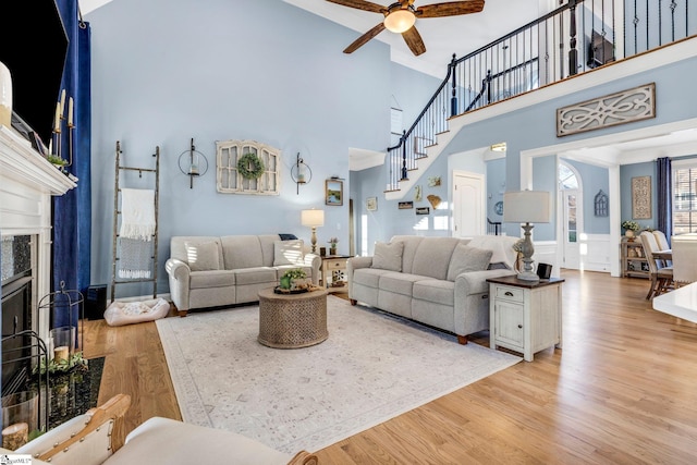 living room with ceiling fan, a fireplace, a towering ceiling, and light wood-type flooring