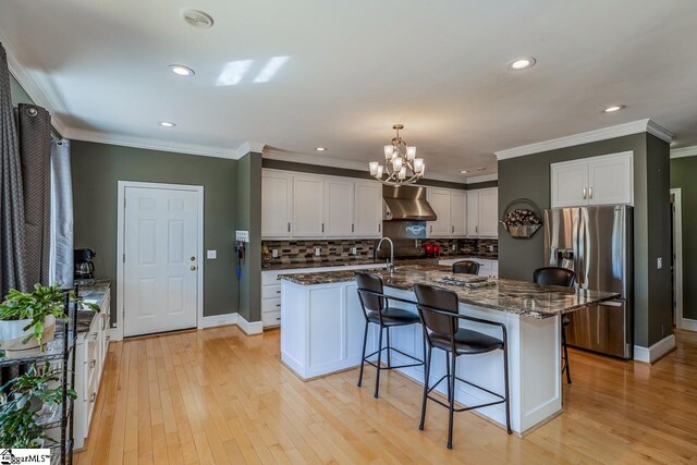 kitchen featuring stainless steel refrigerator with ice dispenser, light wood-style floors, a kitchen island with sink, ventilation hood, and dark stone counters