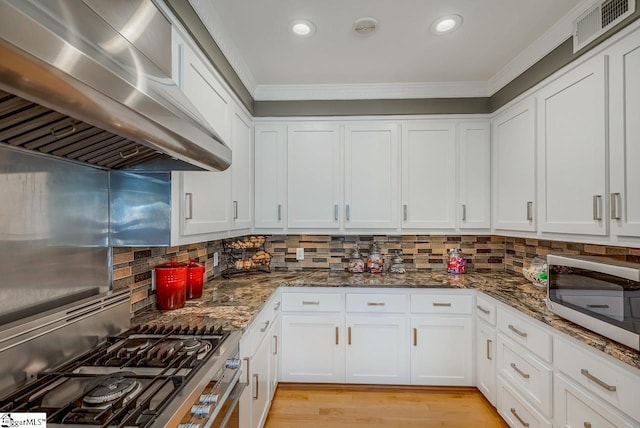 kitchen featuring stainless steel appliances, dark stone counters, white cabinets, exhaust hood, and ornamental molding