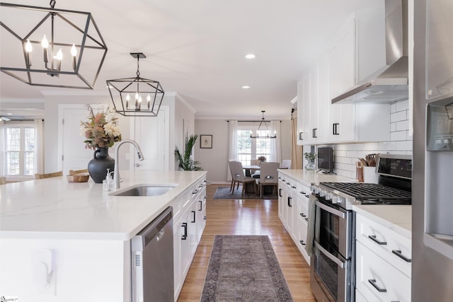 kitchen with a center island with sink, wall chimney range hood, sink, appliances with stainless steel finishes, and decorative light fixtures