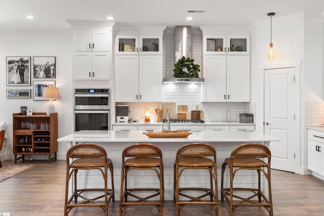 kitchen with decorative light fixtures, wall chimney range hood, an island with sink, and appliances with stainless steel finishes