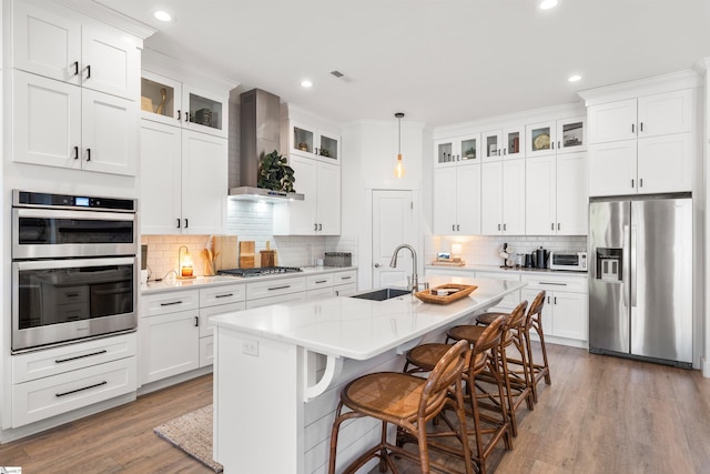 kitchen featuring appliances with stainless steel finishes, sink, wall chimney range hood, pendant lighting, and a center island with sink