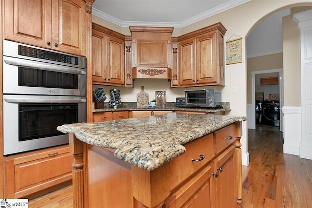 kitchen with washing machine and clothes dryer, stainless steel double oven, light stone counters, crown molding, and a kitchen island