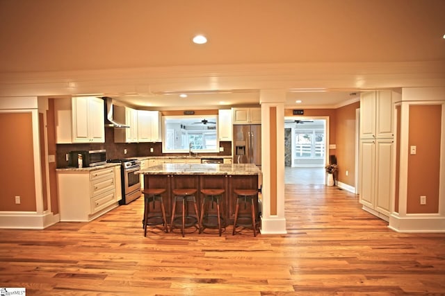 kitchen featuring a center island, wall chimney exhaust hood, stainless steel appliances, a kitchen breakfast bar, and light stone counters