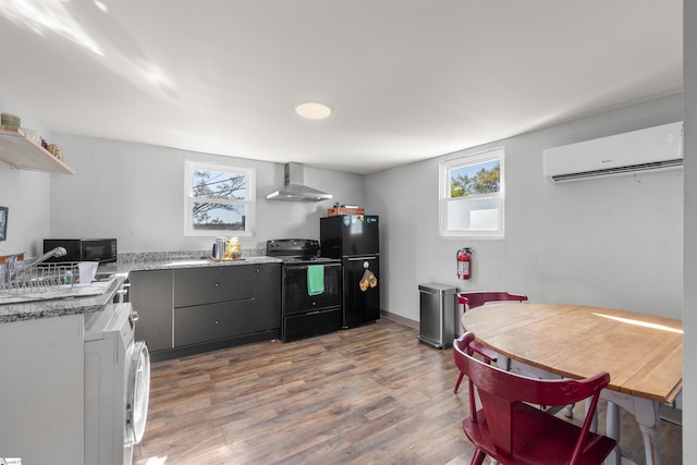 kitchen with black appliances, wall chimney range hood, washer / dryer, wood-type flooring, and a wall unit AC