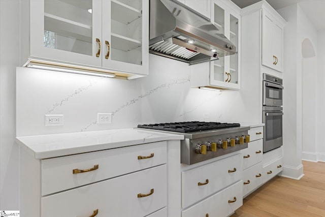 kitchen with white cabinets, appliances with stainless steel finishes, range hood, light hardwood / wood-style floors, and light stone counters