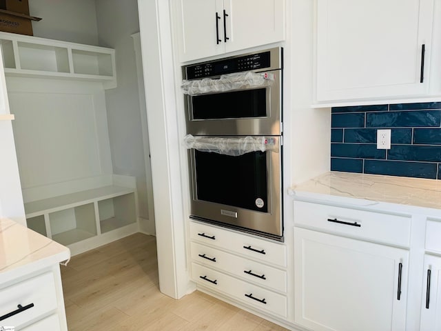 kitchen with white cabinetry, light stone countertops, stainless steel double oven, light hardwood / wood-style flooring, and backsplash