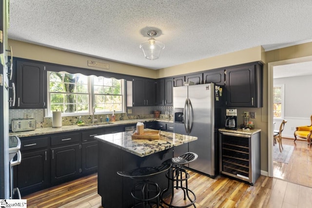 kitchen with light stone countertops, wine cooler, light hardwood / wood-style flooring, stainless steel refrigerator with ice dispenser, and a kitchen island