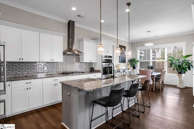 kitchen featuring a kitchen island with sink, sink, wall chimney range hood, pendant lighting, and white cabinetry