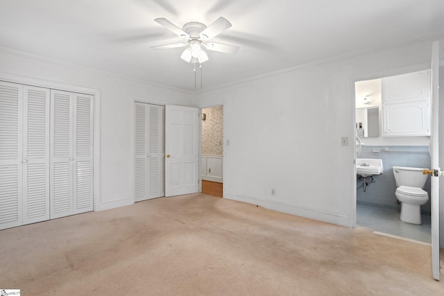 unfurnished bedroom with light carpet, ensuite bath, ceiling fan, and multiple closets